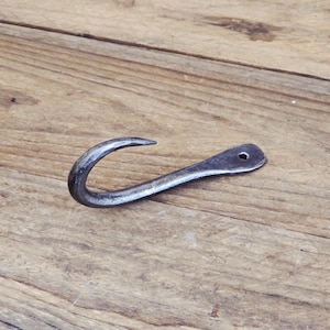 May include: A hand-forged metal hook with a curved design and a small hole at the end. The hook is dark gray and appears to be made of iron or steel. It is designed for hanging items and is shown on a wooden surface.