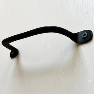 Puede incluir: Un tirador de mueble de metal negro mate con un diseño curvo y arqueado. El tirador presenta dos extremos de montaje planos, cada uno asegurado con un tornillo de cabeza Phillips visible, proporcionando un agarre para puertas o cajones.