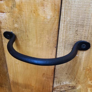 May include: Black metal door handle with a curved design. The handle is attached to a wooden door with screws.