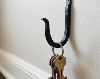 May include: A black metal wall hook with a textured, hand-forged appearance, secured to the wall with a single screw. A silver-toned metal keyring hangs from the hook, holding several brass-colored keys, including one clearly marked with the text Kwikset.