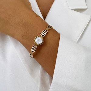 May include: Gold bracelet with a white mother-of-pearl dial featuring Roman numerals. The bracelet has a geometric design.