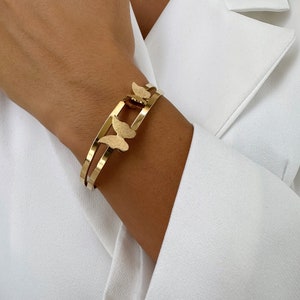 May include: Gold-toned bracelet featuring a butterfly design. The bracelet has a double-band design with two butterflies. The butterflies have a textured finish. The bracelet is worn on a wrist, against a white background.