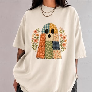 Op de afbeelding: Een off-white t-shirt met korte mouwen en een centrale afbeelding van een geest gemaakt van verschillende patchwork-stofpatronen, waaronder blauwe ruitjes, oranje bloemen en groene bloemen, geflankeerd door twee stengels met oranje bloemen.