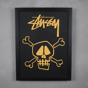 May include: A black framed print with a gold skull and crossbones design. The text "STUSSY" is printed in gold above the skull.