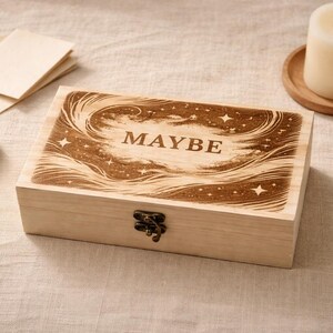 May include: A wooden box with the word "MAYBE" engraved on the lid, featuring a celestial design. The box is closed with a metal clasp. A lit candle sits on a wooden coaster, and a small stone and notecards are nearby.