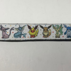 Könnte beinhalten: Ein weißes Schlüsselband mit bunten Cartoonfiguren. Das Band hat einen schwarzen Rand und einen silbernen Schlüsselring. Die Figuren sind in einer Reihe angeordnet, mit einem Tupfenmuster.