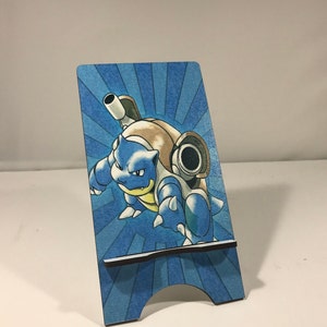 May include: A blue and brown Pokemon phone stand featuring Blastoise, a water-type Pokemon. The stand has a blue and white striped background.