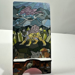 May include: A stained glass style phone stand featuring three Pokemon: a blue and purple legendary Pokemon, a yellow and brown legendary Pokemon, and a brown and orange legendary Pokemon.