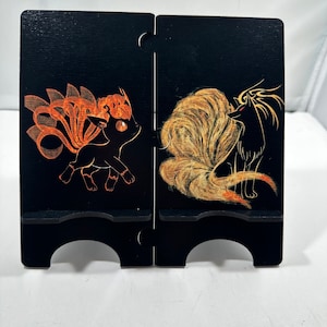 May include: Black wooden phone stand with a painted image of two fox-like creatures. The creature on the left is orange and the creature on the right is black and white.