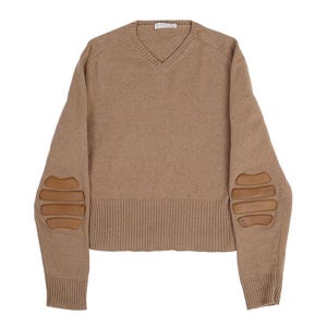 Puede incluir: Un jersey beige con cuello en V y coderas marrones. El jersey tiene puños y dobladillo acanalados. Las coderas son rectangulares y están cosidas a las mangas. El jersey está hecho de un material suave.