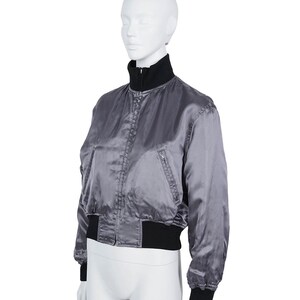 May include: A grey satin bomber jacket with a black collar, cuffs, and waistband. The jacket has a zip closure and a small pocket on the left side. The jacket is displayed on a white mannequin.
