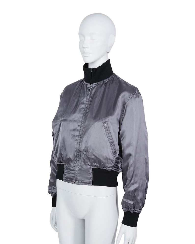 May include: A grey satin bomber jacket with a black collar, cuffs, and waistband. The jacket has a zip closure and a small pocket on the left side. The jacket is displayed on a white mannequin.