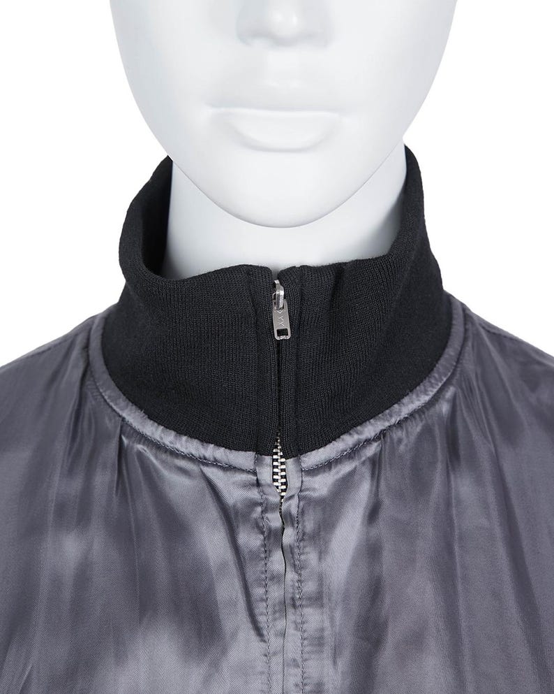 May include: A close-up of a grey bomber jacket with a black ribbed collar. The jacket has a silver zip running down the front. The jacket is displayed on a mannequin.