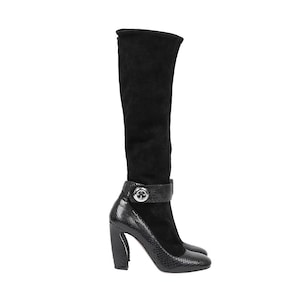 May include: A black knee-high boot featuring a soft suede upper and a textured snakeskin-patterned leather lower section. The boot includes a decorative ankle strap secured with a prominent, circular silver-toned metal button detail and a block heel.