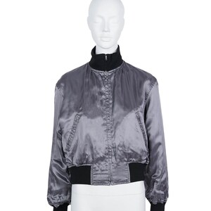 May include: A grey satin bomber jacket with a black collar, cuffs, and hem. The jacket has a zip closure and two front pockets. The jacket is displayed on a mannequin.