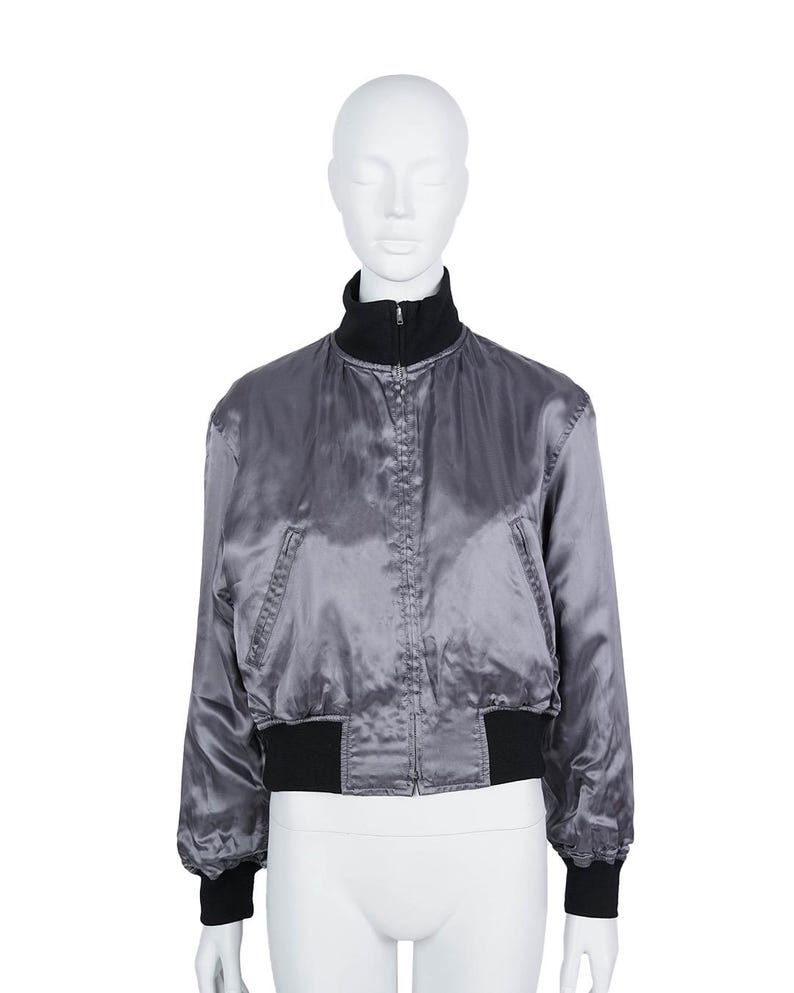 May include: A grey satin bomber jacket with a black collar, cuffs, and hem. The jacket has a zip closure and two front pockets. The jacket is displayed on a mannequin.