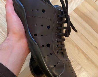 May include: Brown leather shoe with a black sole and laces. The shoe features multiple small circular perforations across the upper. The laces are dark brown. The shoe is held in a person's hand.