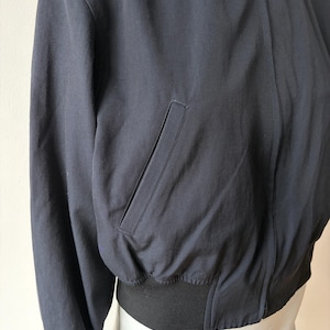 May include: Navy blue bomber jacket with a black ribbed hem and cuffs. The jacket features a front zip closure and a single pocket on the front. The jacket is made of a smooth, matte fabric.