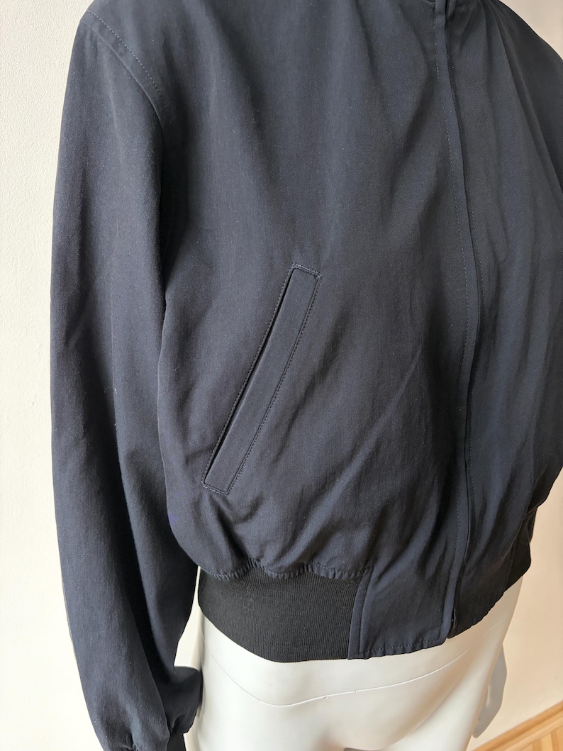 May include: Navy blue bomber jacket with a black ribbed hem and cuffs. The jacket features a front zip closure and a single pocket on the front. The jacket is made of a smooth, matte fabric.
