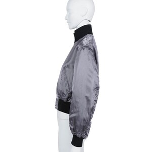 May include: A grey satin bomber jacket with a black collar and cuffs. The jacket features a zip-up front and a slightly puffed sleeve design. The jacket is displayed on a mannequin.