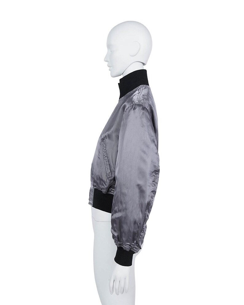 May include: A grey satin bomber jacket with a black collar and cuffs. The jacket features a zip-up front and a slightly puffed sleeve design. The jacket is displayed on a mannequin.
