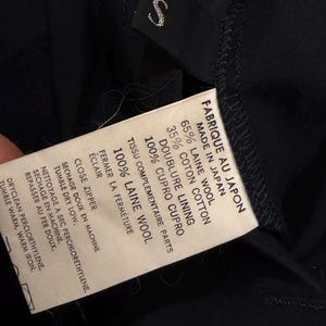 May include: A close-up of a black garment label with white text. The label states "FABRIQUE AU JAPON MADE IN JAPAN" and details fabric content: 65% wool, 35% cotton, and 100% cupro lining. Washing instructions are also included.