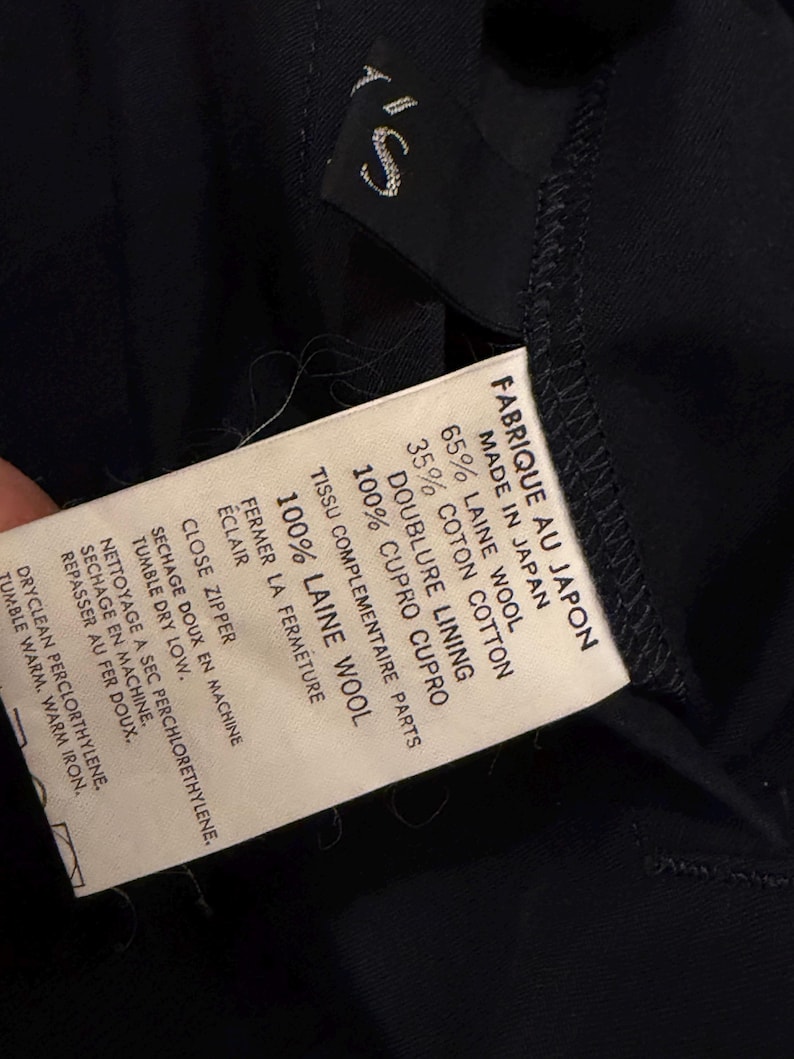 May include: A close-up of a black garment label with white text. The label states "FABRIQUE AU JAPON MADE IN JAPAN" and details fabric content: 65% wool, 35% cotton, and 100% cupro lining. Washing instructions are also included.
