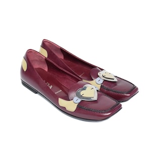 Könnte beinhalten: Ein Paar burgunderfarbene Lederschuhe mit einem herzförmigen Metallakzent. Die Schuhe haben eine quadratische Spitze und einen niedrigen Absatz. Der Herz-Akzent ist silber mit goldener Einfassung und einem gelben Herzen in der Mitte. Die Schuhe sind in Italien hergestellt.