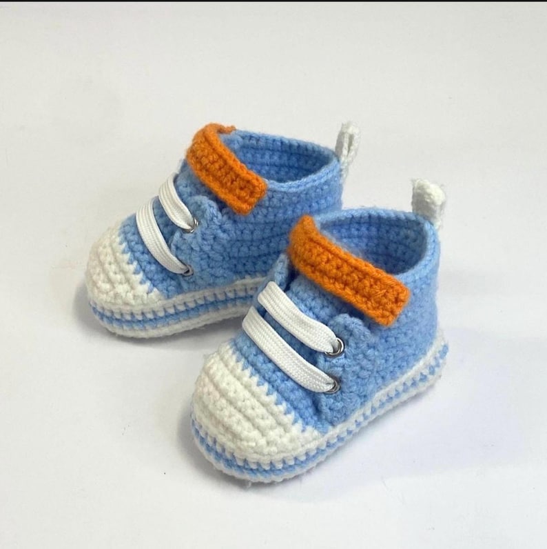 May include: A pair of handmade baby sneakers. The crochet shoes are light blue with white soles and toe caps. They have orange straps and white laces. The shoes are designed to look like miniature high-top sneakers.