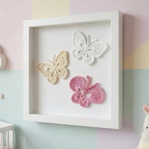 May include: A white framed wall art piece featuring three crocheted butterflies. The butterflies are in shades of cream, white, and pink. The frame is square and white, creating a clean and modern look. The butterflies are arranged on a white background.