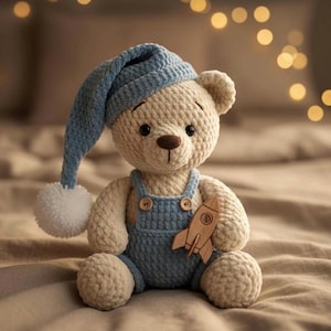 May include: A handmade teddy bear wearing a blue knit nightcap with a white pom-pom and blue overalls. The bear holds a wooden rocket and sits on a bed. The bear is beige with brown eyes.