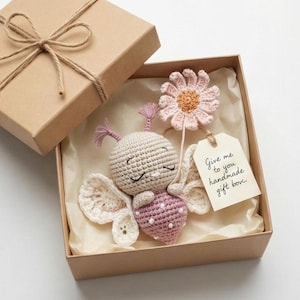 Può includere: Un giocattolo a farfalla fatto a mano all'uncinetto con corpo rosa e ali color crema, presentato in una scatola regalo marrone. La scatola è legata con spago e contiene un'etichetta con la scritta "Give me to you handmade gift box."