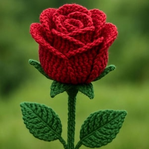 May include: A handcrafted, red crocheted rose with green leaves and stem. The rose is in full bloom, showcasing detailed petal work. The leaves and stem are a vibrant green, contrasting with the deep red of the flower. The background is a soft, blurred green.