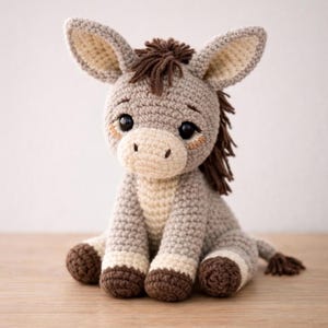 May include: A handmade crochet donkey plush toy in shades of gray, cream, and brown. The donkey has large, expressive eyes, a cream-colored muzzle, and a brown mane and tail. The plush is sitting on a wooden surface.