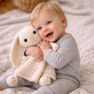 Puede incluir: Un bebé sonriente con pijama gris abraza un conejito de ganchillo color crema. El conejito tiene orejas largas, ojos negros de botón y un cuerpo texturizado. El fondo es una alfombra suave de tonos neutros.