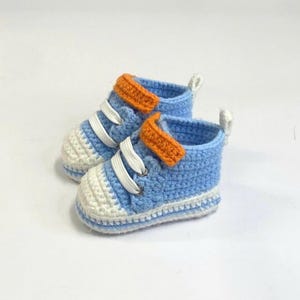 May include: A pair of baby crochet sneakers in light blue, white, and orange. The shoes feature white laces, a white toe cap, and an orange strap across the top. The sneakers are designed for infants.
