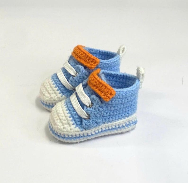 May include: A pair of baby crochet sneakers in light blue, white, and orange. The shoes feature white laces, a white toe cap, and an orange strap across the top. The sneakers are designed for infants.