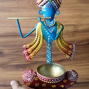 May include: A colorful metal figurine of a figure playing a flute, with a blue body, yellow and red accents, and a decorative headdress. The figurine has a small gold bowl at the base and is held by a hand.