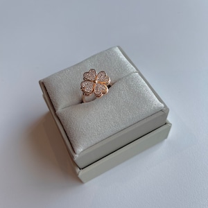 May include: A rose gold ring featuring a four-leaf clover design. Each leaf is shaped like a heart and encrusted with small, clear crystals. The ring is displayed in a gray jewelry box.