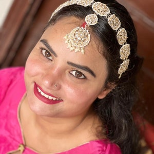 May include: A woman wearing a pink top with a gold and white jeweled headband and a matching forehead ornament.