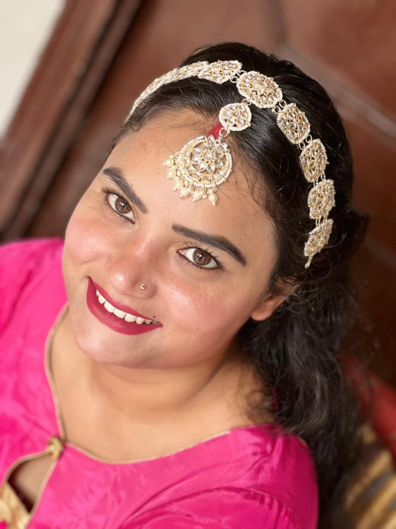 May include: A woman wearing a pink top with a gold and white jeweled headband and a matching forehead ornament.