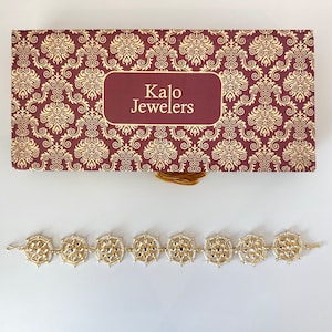May include: A gold bracelet with white beads and a decorative design. The bracelet is in a box with the words "Kalo Jewelers" on it.