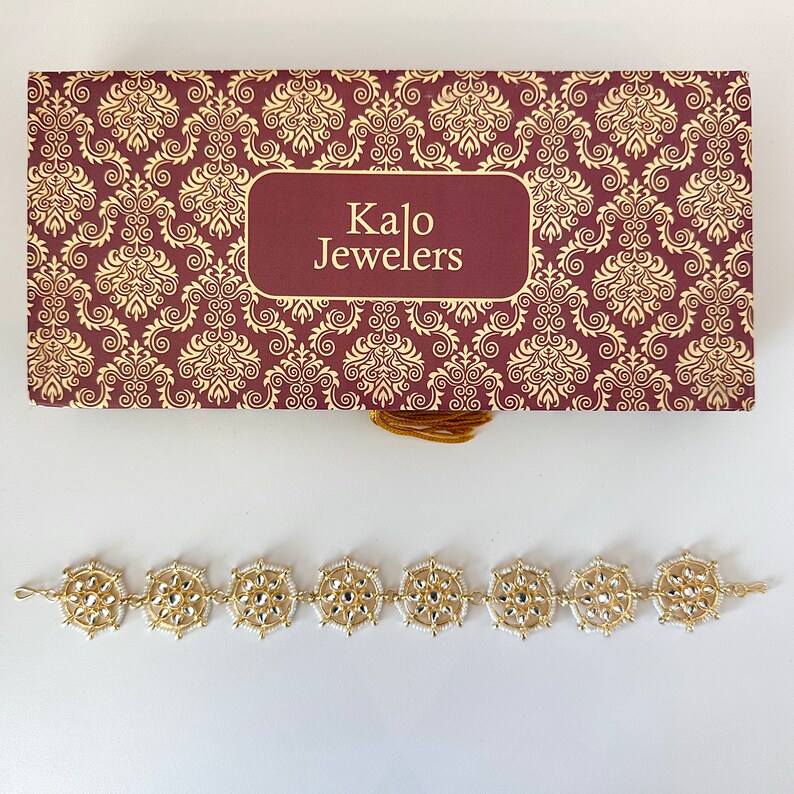 May include: A gold bracelet with white beads and a decorative design. The bracelet is in a box with the words "Kalo Jewelers" on it.