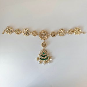 May include: A gold and white headpiece with a green and gold pendant. The headpiece is made of small round pieces connected by a chain. The pendant is shaped like a crescent moon with a green stone in the centre.