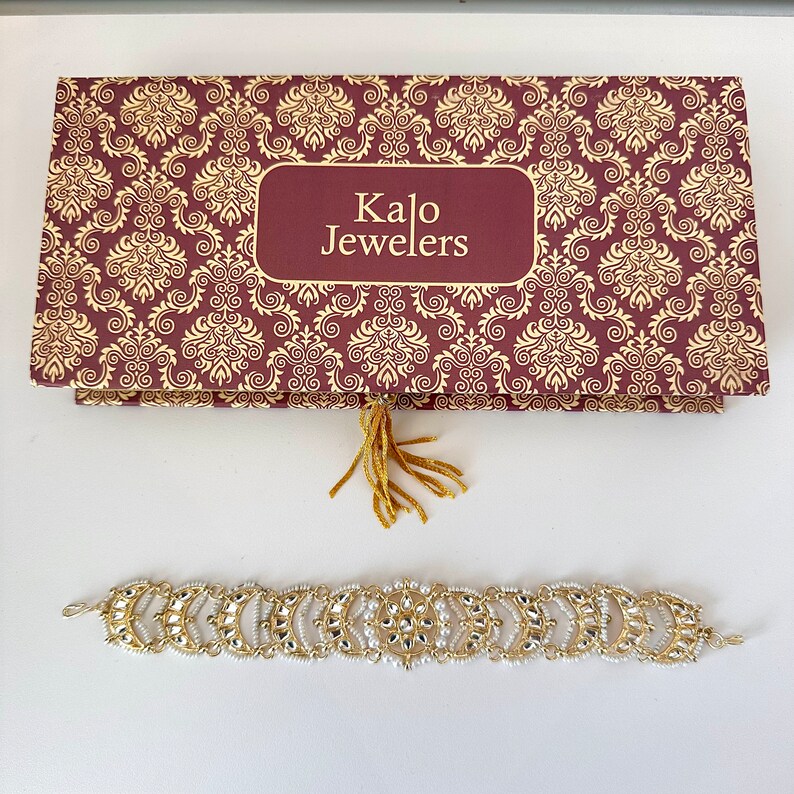 May include: A gold and white beaded bracelet with a decorative design. The bracelet is in a box with the words "Kalo Jewelers" on it.