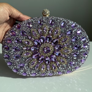 May include: Oval-shaped clutch covered in silver and purple rhinestones. The center features a floral design with purple and gold-toned gems. A small, round, gold-toned clasp is at the top.