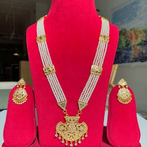 May include: A gold and white pearl necklace set with a large, ornate pendant and matching earrings. The necklace has five strands of pearls and the pendant is shaped like a fan with intricate details.