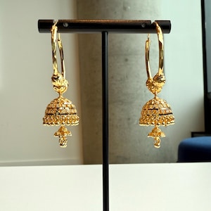May include: A pair of gold-colored earrings with a bell-shaped design. The earrings are suspended from a hoop and feature intricate detailing, including a floral-like pattern at the top and a tiered bell shape with a small, dangling element at the bottom. The earrings are displayed on a black stand.