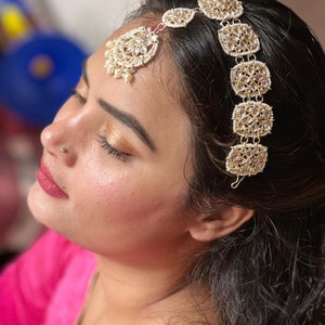 May include: A woman wearing a pink top with a gold and white jeweled headpiece. The headpiece has a teardrop-shaped pendant with pearls and a row of square-shaped ornaments.