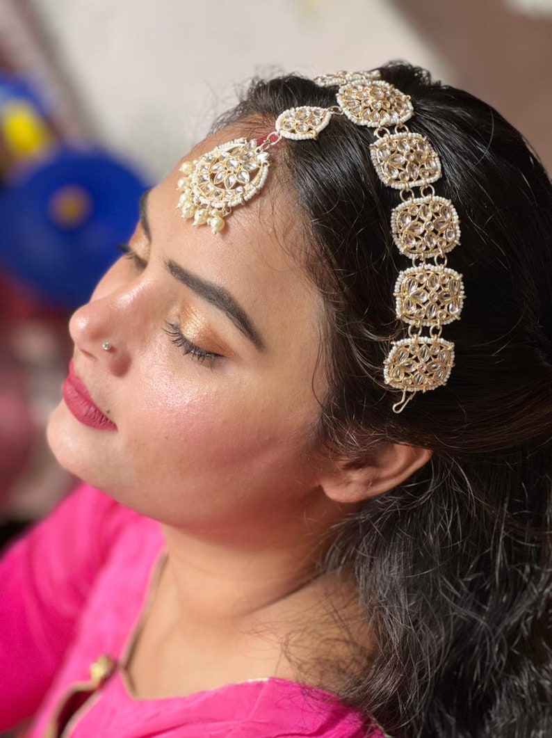 May include: A woman wearing a pink top with a gold and white jeweled headpiece. The headpiece has a teardrop-shaped pendant with pearls and a row of square-shaped ornaments.